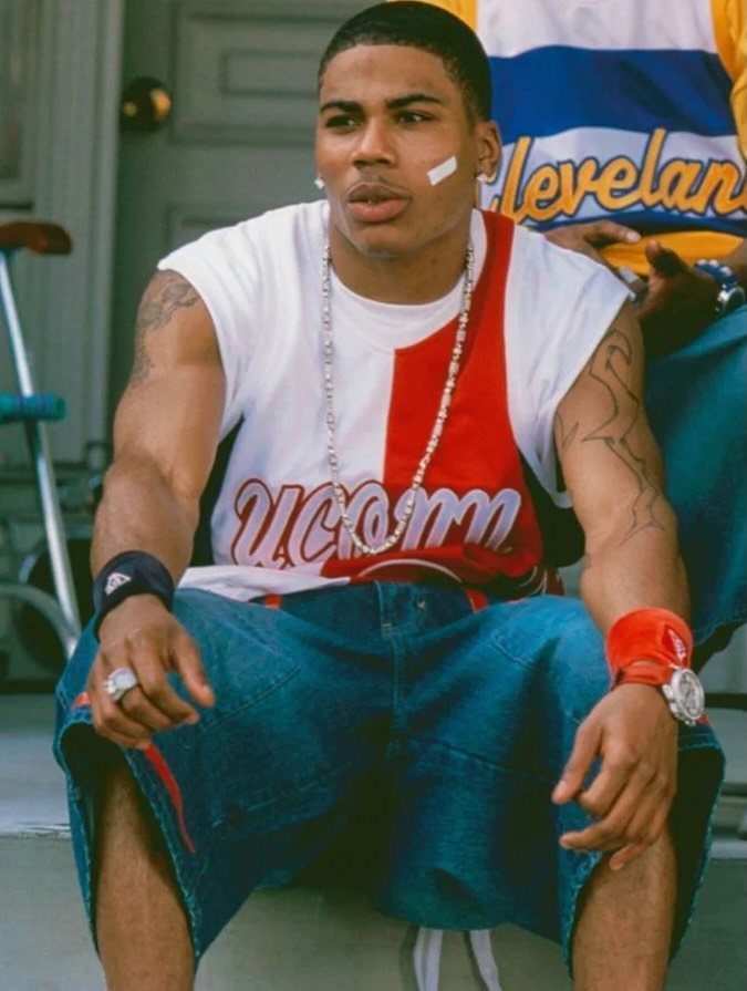 Young man in baggy basketball jersey and denim shorts sitting outdoors wearing silver jewelry.