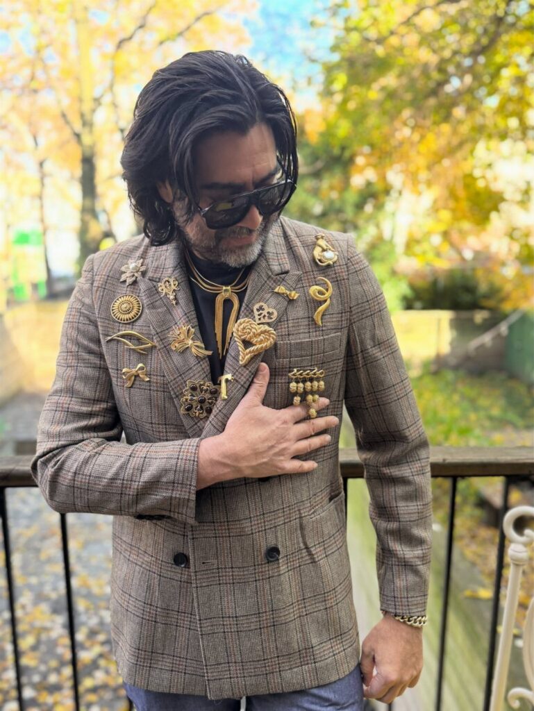 Man wearing a plaid blazer covered in many large, ornate gold decorative brooches.