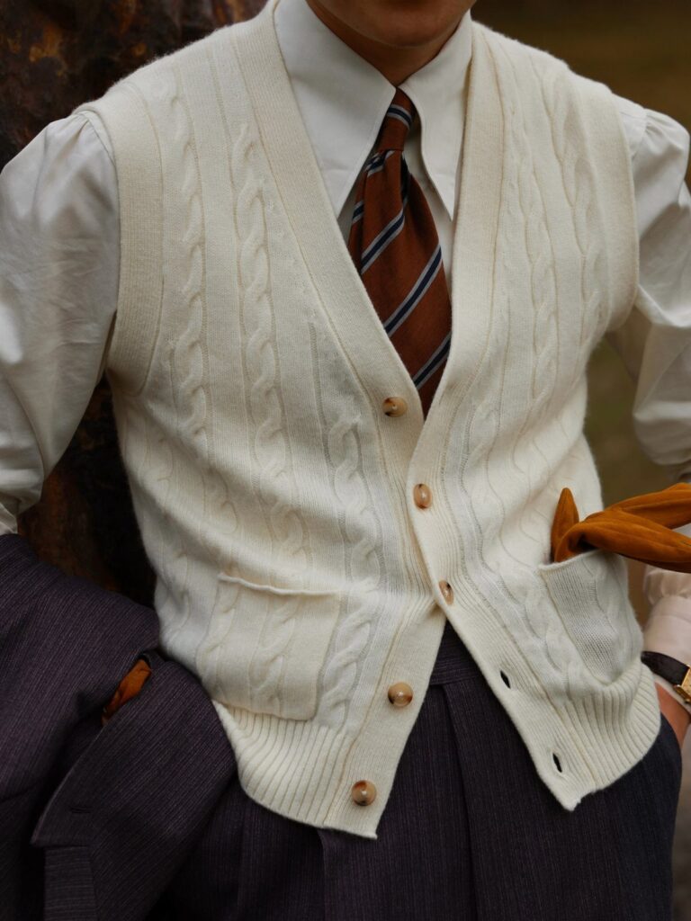 A person wearing a cream cable-knit sweater vest, white shirt, and striped tie.
