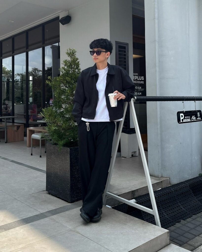 A person in a black jacket, white t-shirt, and black trousers standing outdoors.