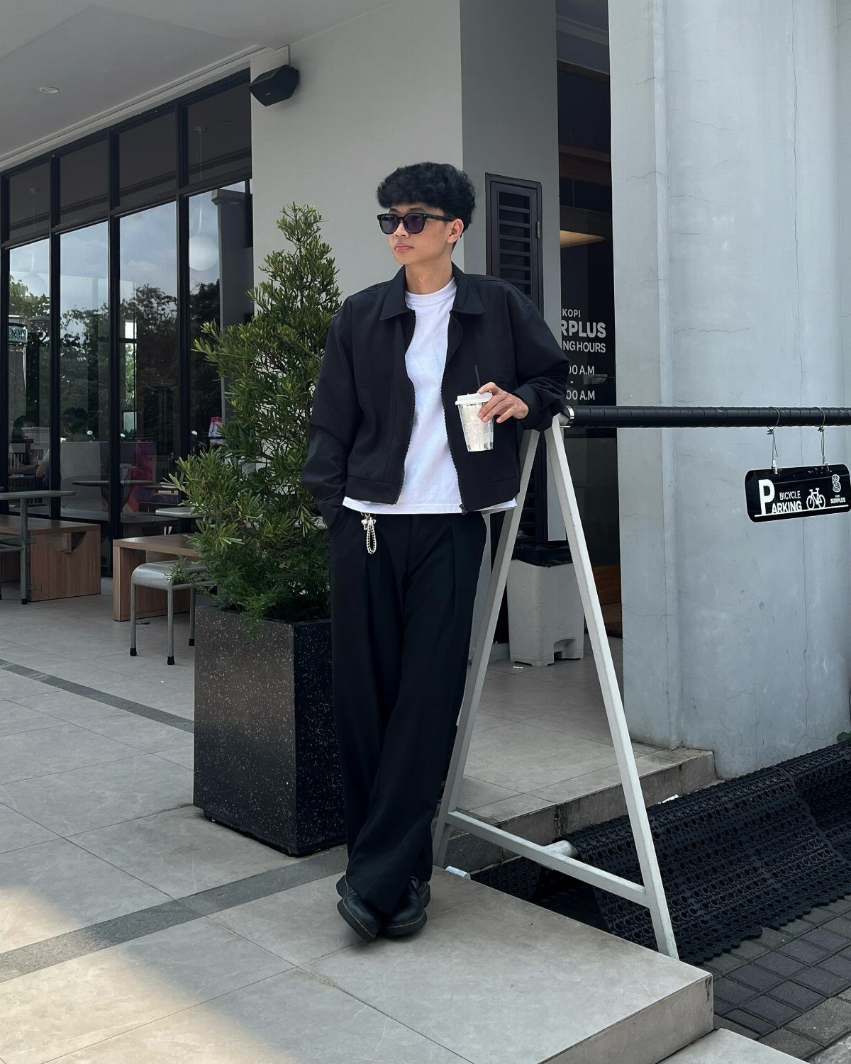 A person in a black jacket, white t-shirt, and black trousers standing outdoors.