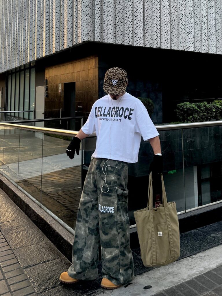 A person wearing a white t-shirt, camouflage pants, and a leopard print hat standing outdoors.