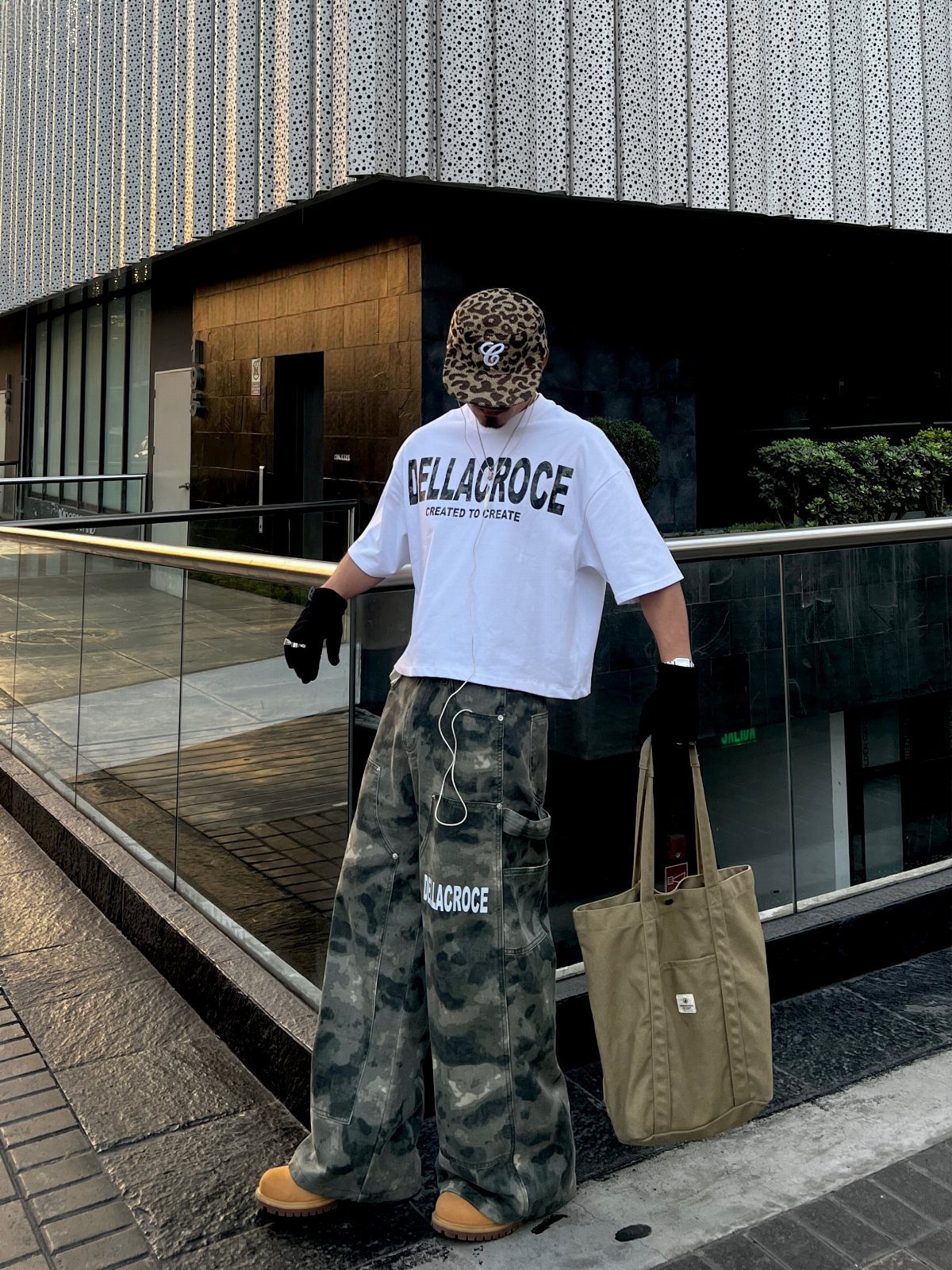 A person wearing a white t-shirt, camouflage pants, and a leopard print hat standing outdoors.