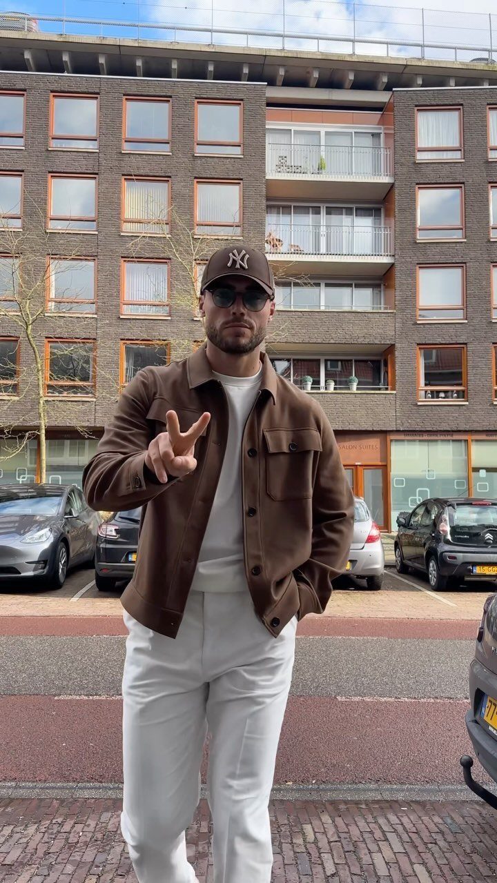 Man posing in a brown jacket, white t-shirt, white trousers, and a brown baseball cap.