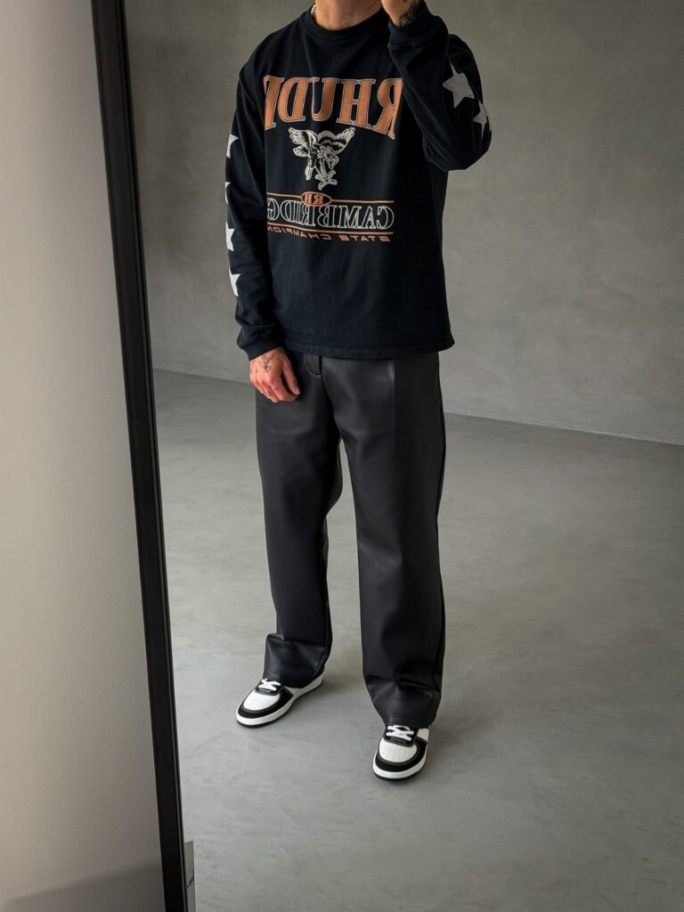 A man wearing a black graphic long-sleeve shirt, black leather pants, and black and white sneakers.