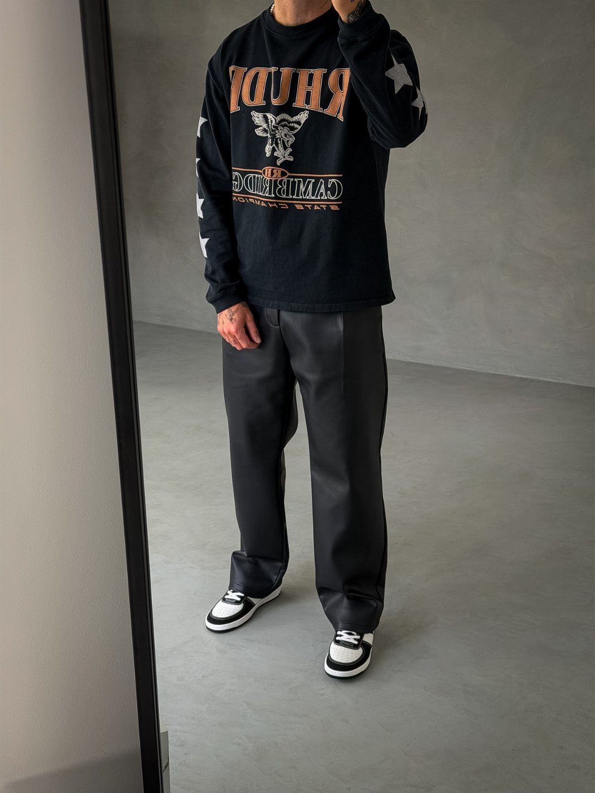 A man wearing a black graphic long-sleeve shirt, black leather pants, and black and white sneakers.