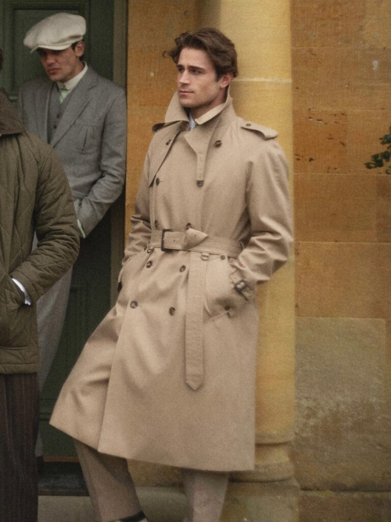 A man wearing a long tan belted trench coat standing against a stone wall.