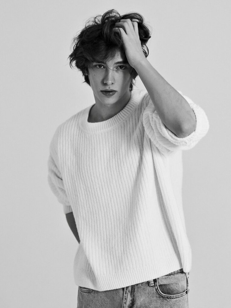 A young man with wavy hair wearing a white ribbed sweater posing against a plain background.