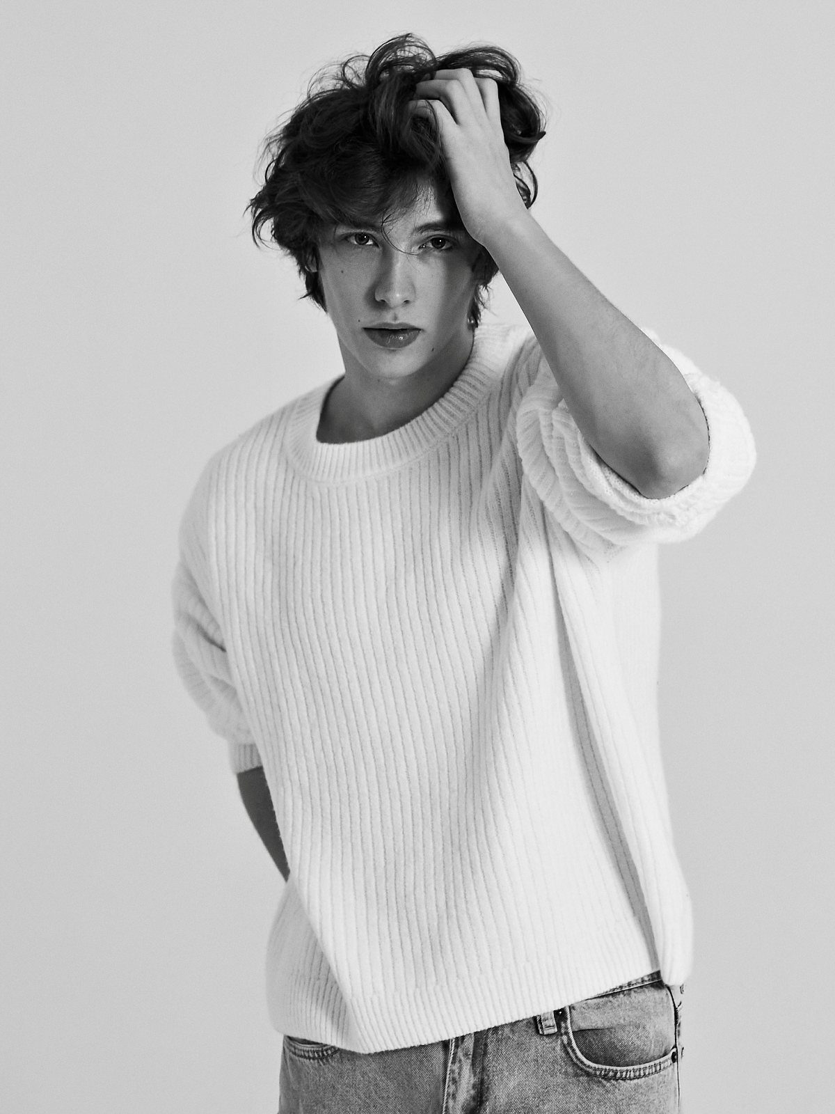A young man with wavy hair wearing a white ribbed sweater posing against a plain background.
