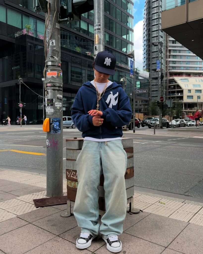 A man in a navy Yankees hoodie, baggy jeans, and a baseball cap standing on a city street.
