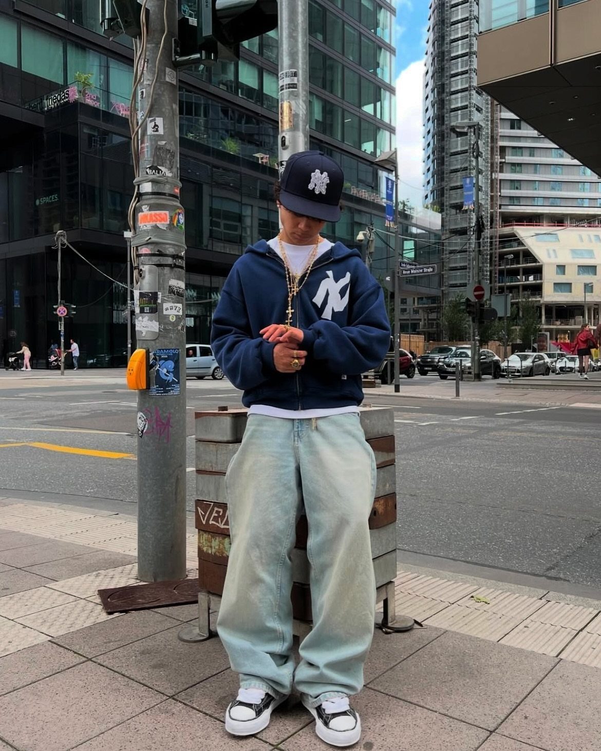 A man in a navy Yankees hoodie, baggy jeans, and a baseball cap standing on a city street.