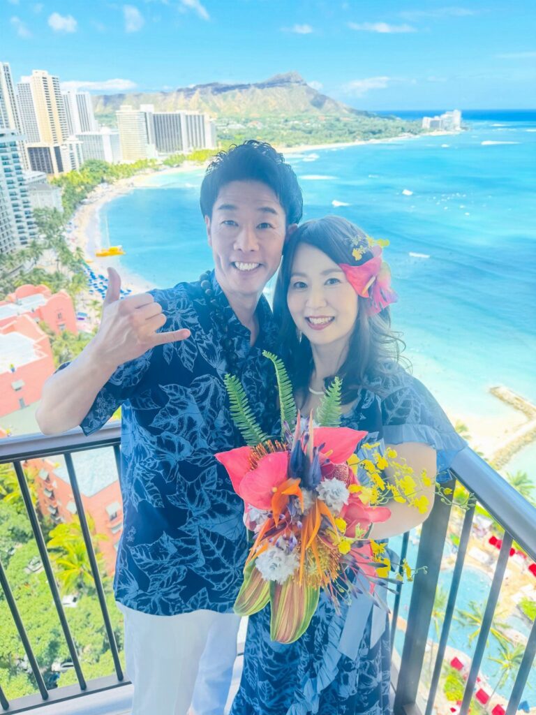 A smiling couple posing on a balcony overlooking a tropical beach with blue ocean water.