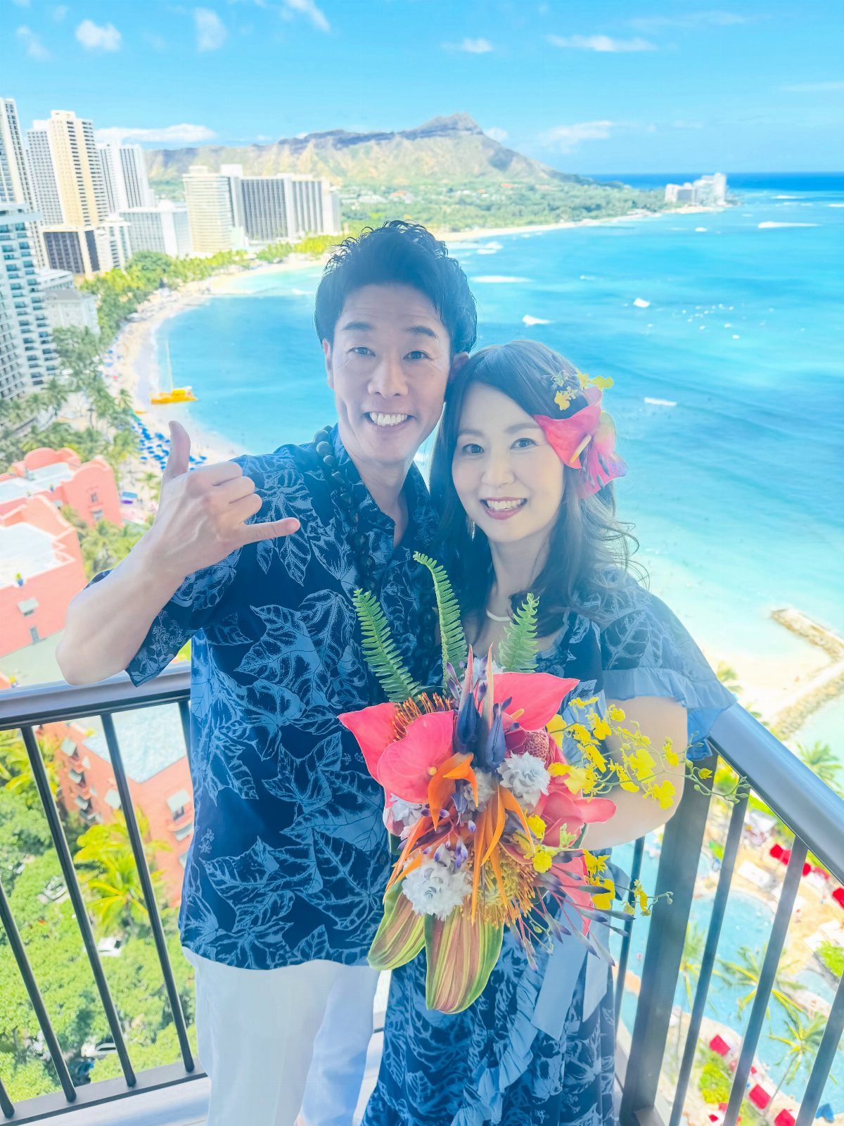 A smiling couple posing on a balcony overlooking a tropical beach with blue ocean water.