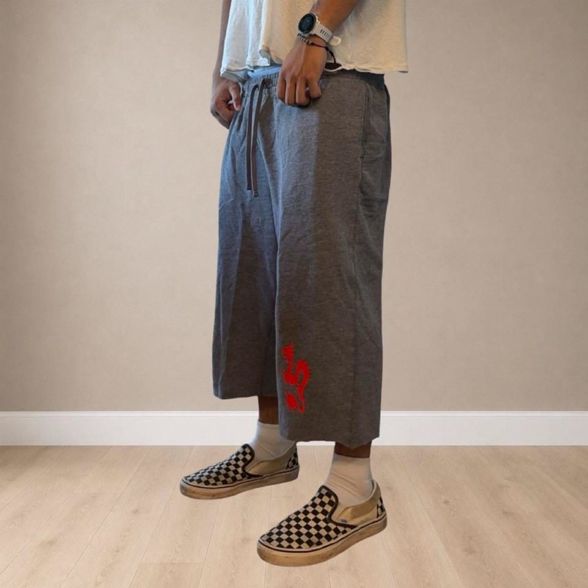 Person wearing grey baggy capri pants, white socks, and checkered slip-on sneakers against a plain wall.
