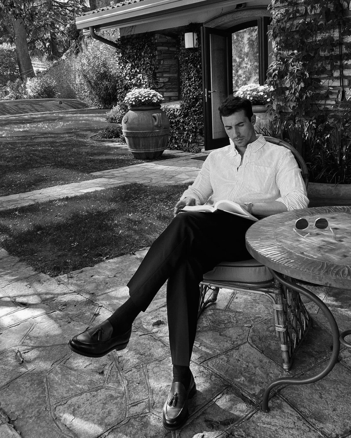 Man in a patterned shirt and dark trousers reading a book on a garden patio.