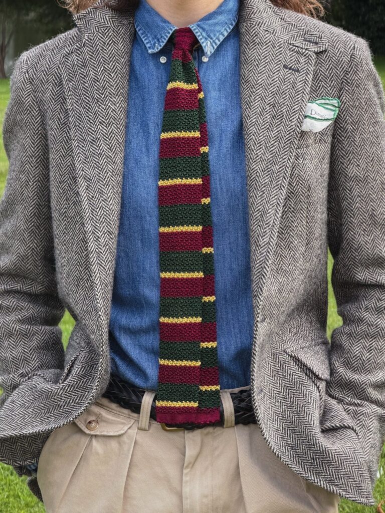 A person dressed in a herringbone blazer, denim shirt, striped knit tie, and corduroy trousers.
