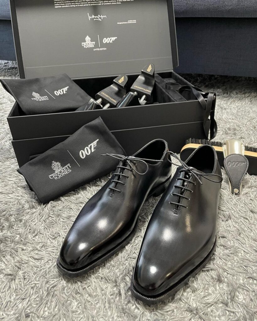 Polished black leather derby shoes displayed with a 007 branded gift box on a grey rug.