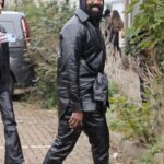 A man in an all-black leather hooded jacket and trousers walking outdoors near green bushes.