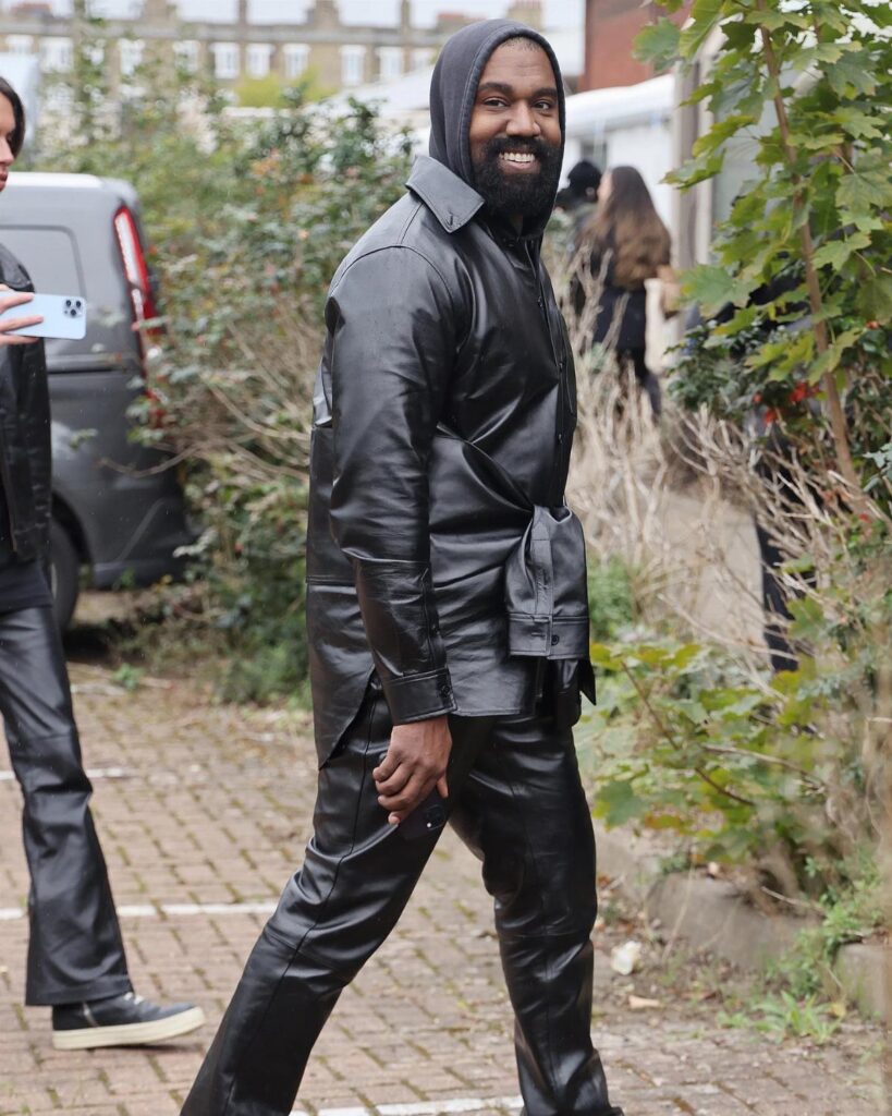A man in an all-black leather hooded jacket and trousers walking outdoors near green bushes.