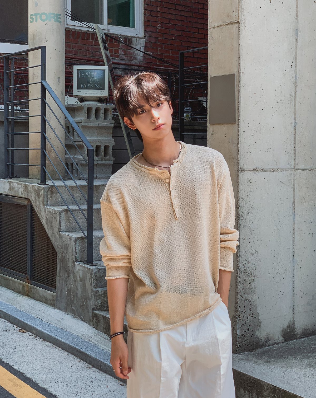 A young man wearing a textured cream henley shirt and white trousers standing outdoors.
