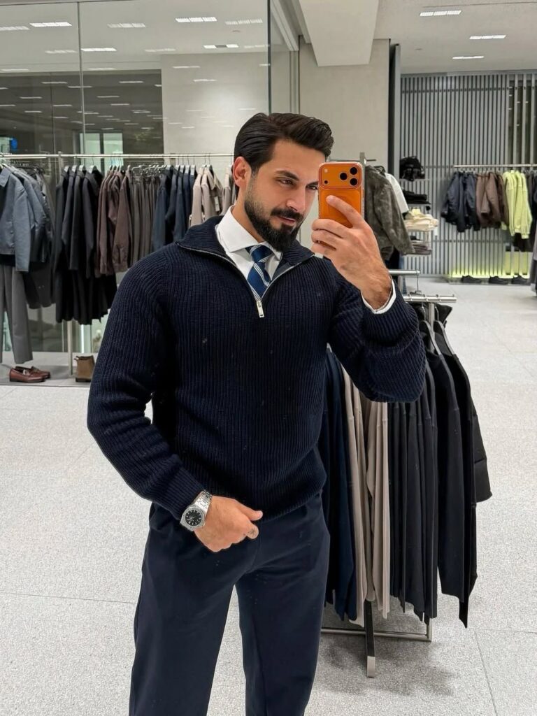 A man posing in a navy blue ribbed quarter-zip sweater, white shirt, and patterned tie.