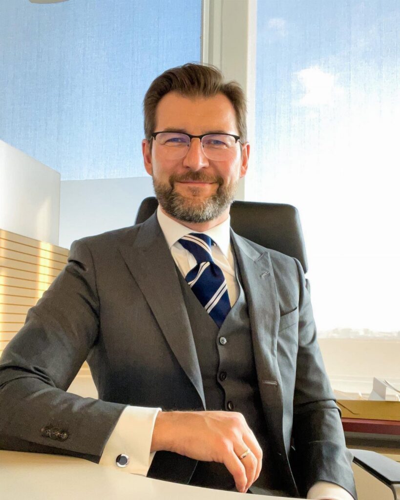 A bearded man in a gray three-piece suit and white shirt sitting at an office desk.