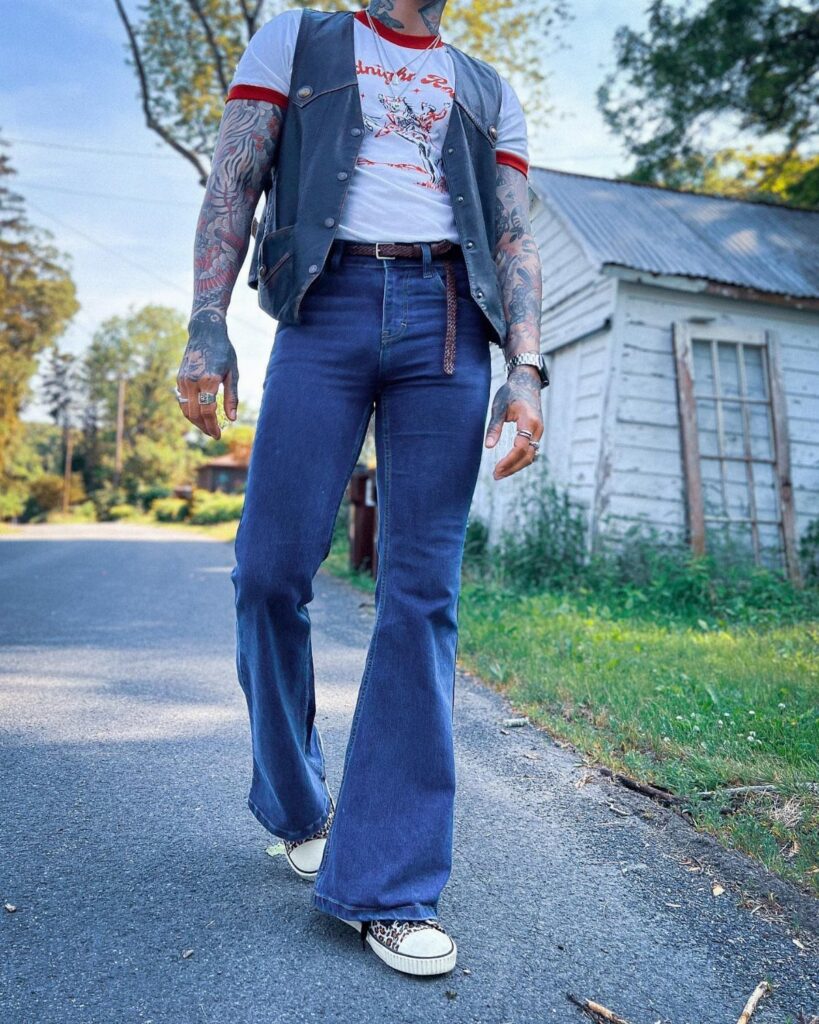 Man wearing blue flared jeans, a white t-shirt, and a dark vest walking outdoors.