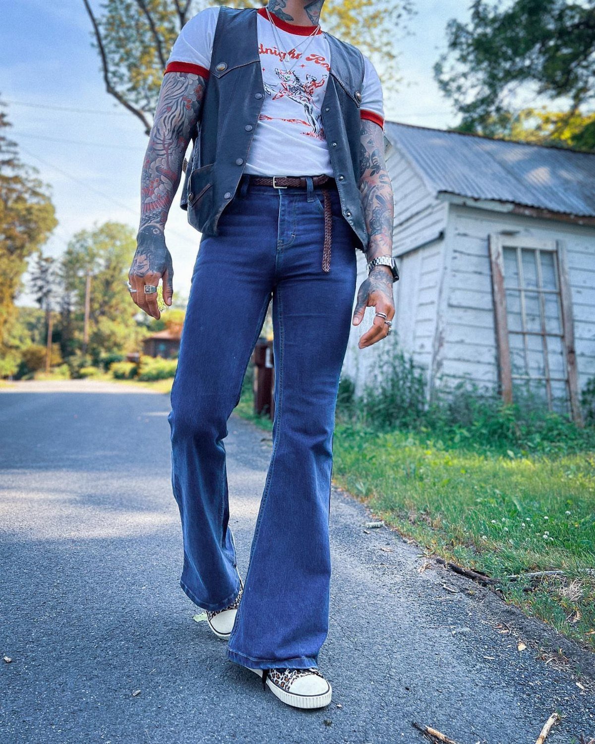 Man wearing blue flared jeans, a white t-shirt, and a dark vest walking outdoors.