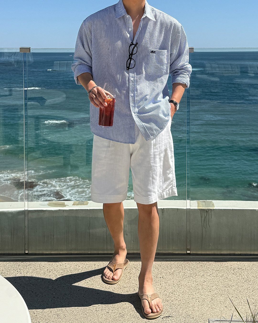 Man in striped seersucker shirt and white linen shorts standing by the ocean.