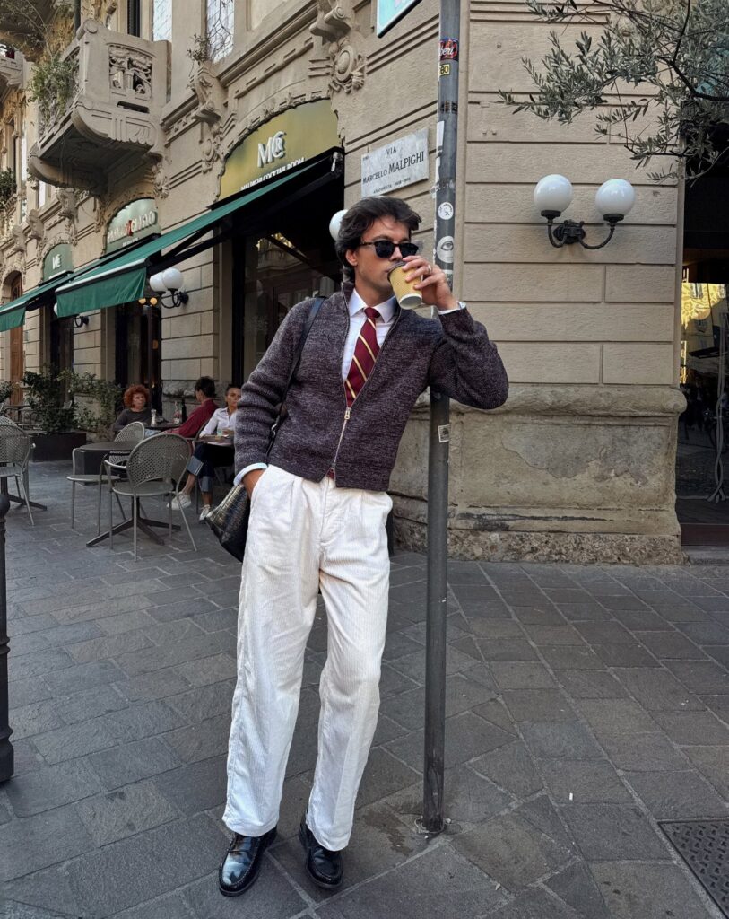 A man wearing a collared shirt, tie, and cardigan layered over white trousers.