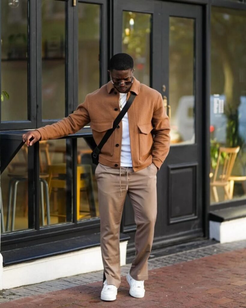 Man wearing a brown jacket, white t-shirt, tan trousers, and white sneakers standing outdoors.