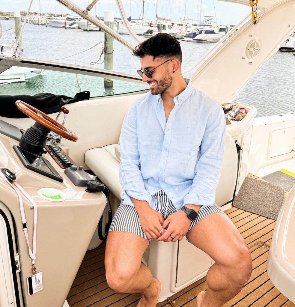 A smiling man in a linen shirt and striped shorts sitting on a boat.
