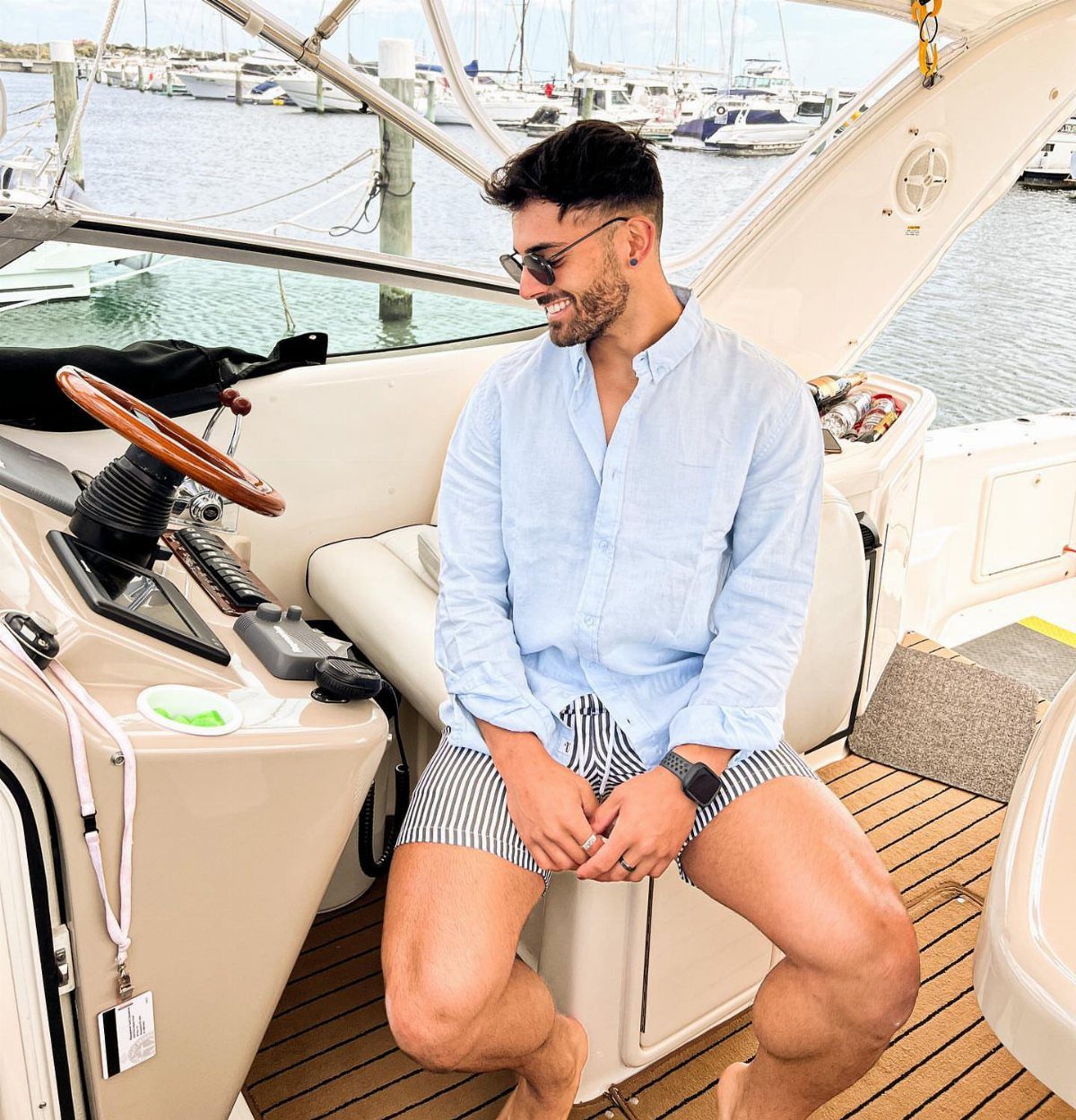 A smiling man in a linen shirt and striped shorts sitting on a boat.