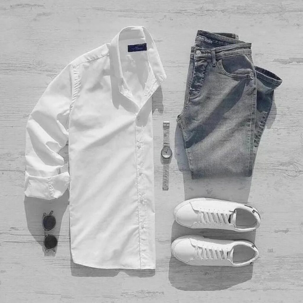 White button-down shirt, blue jeans, white sneakers, sunglasses, and a watch laid out on wood.
