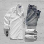 White button-down shirt, blue jeans, white sneakers, sunglasses, and a watch laid out on wood.
