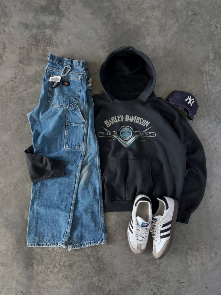 Flat lay of blue denim jeans, black Harley-Davidson hoodie, white sneakers, and a navy baseball cap.