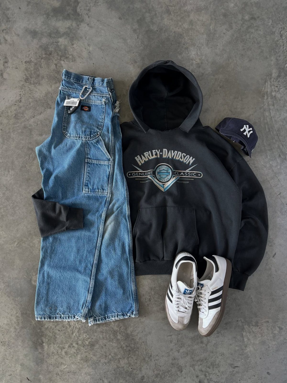 Flat lay of blue denim jeans, black Harley-Davidson hoodie, white sneakers, and a navy baseball cap.