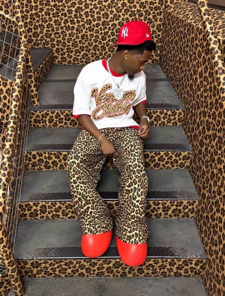 Man sitting on leopard print stairs wearing a white graphic tee and leopard print pants.