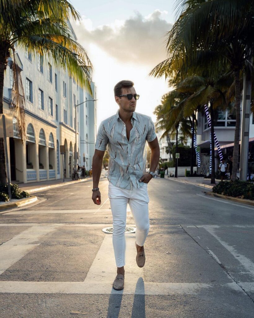 A man in a patterned shirt and white pants walking down a palm-lined street.