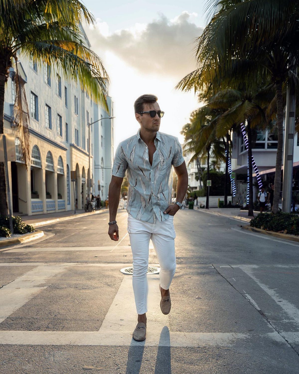A man in a patterned shirt and white pants walking down a palm-lined street.