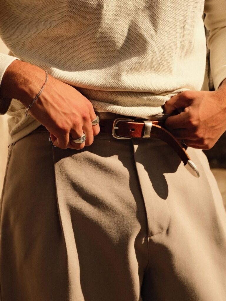 Person wearing a cream knit top, beige trousers, silver rings, and a brown leather belt.