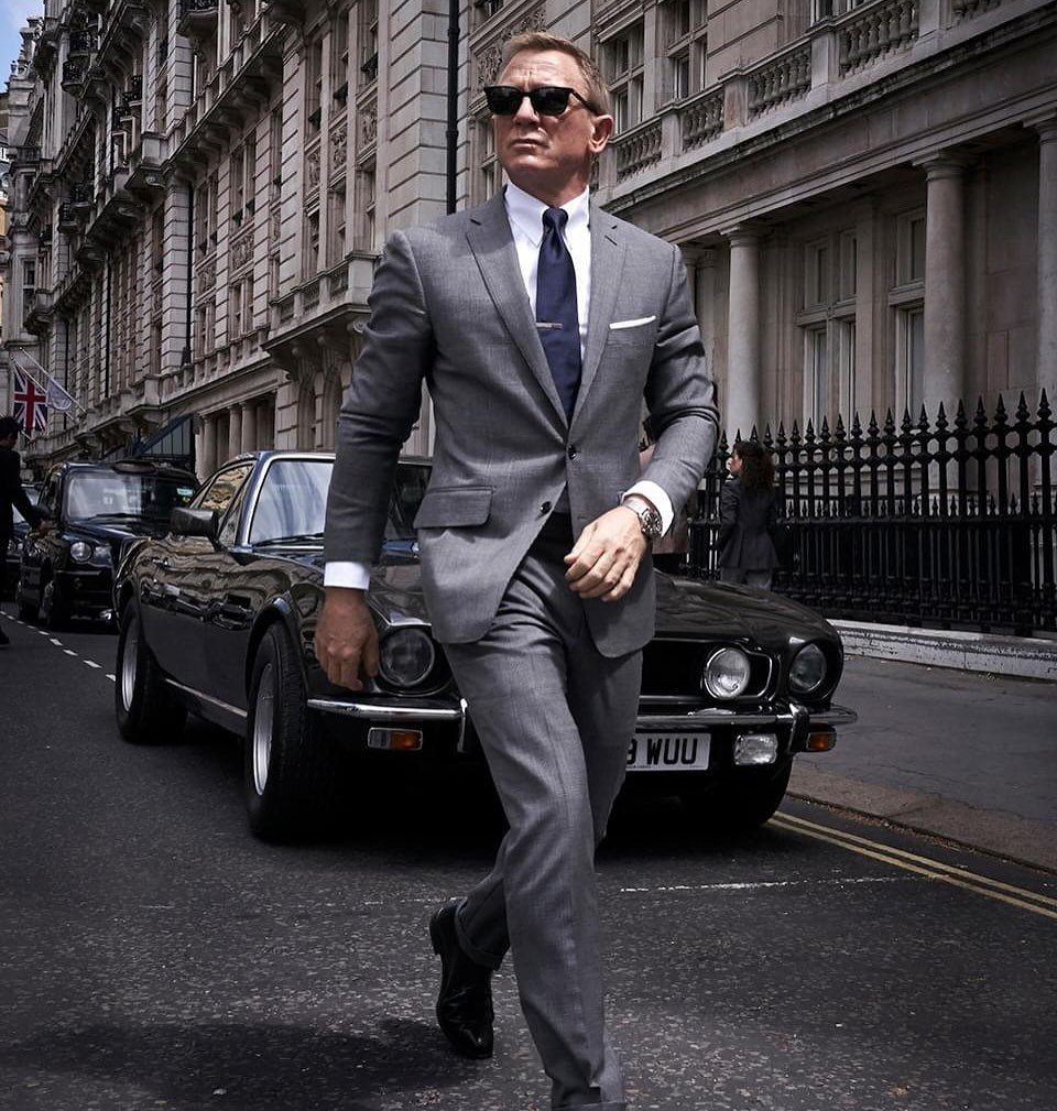 Man in a grey tailored suit and sunglasses walking past a vintage black car.