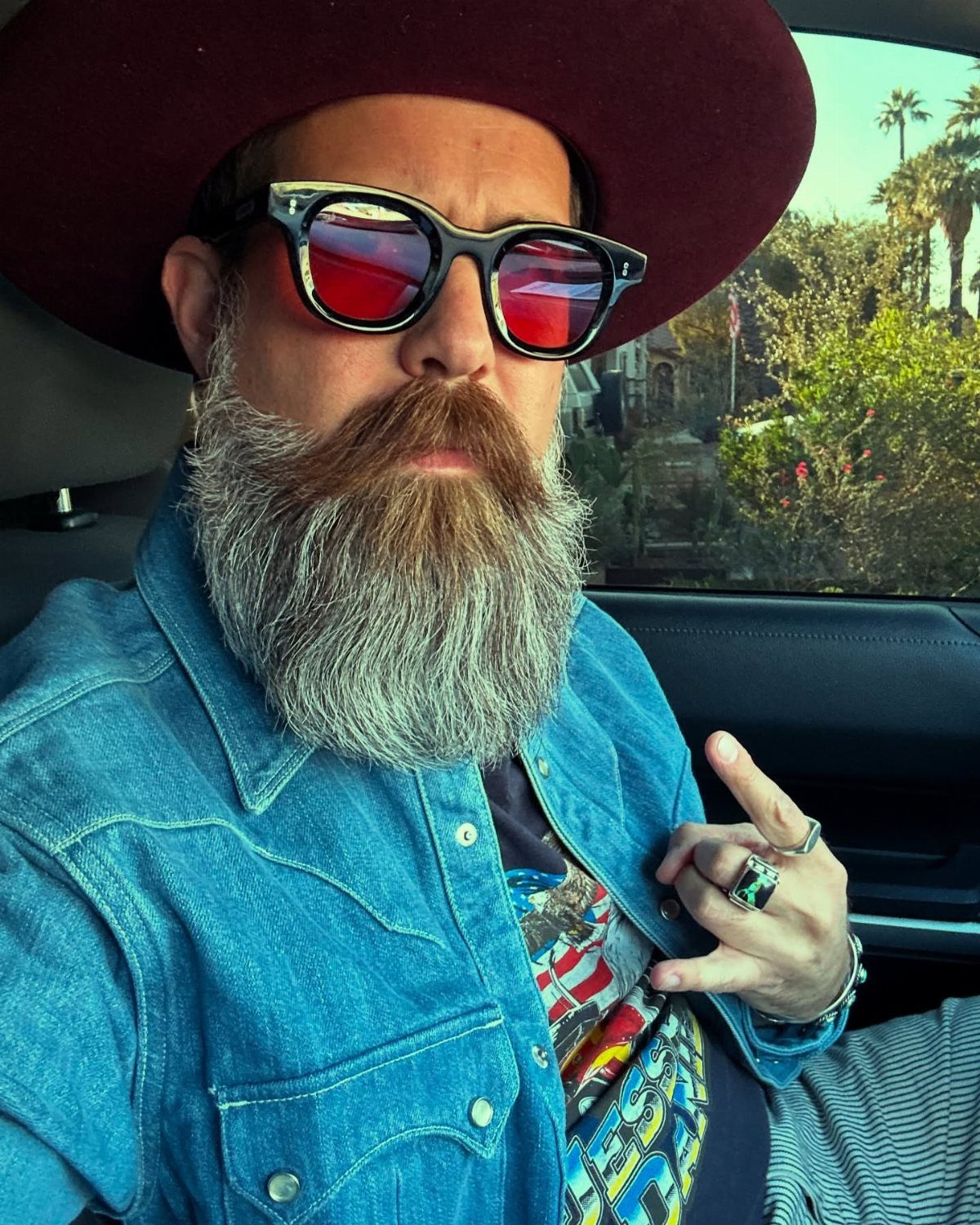 A bearded man wearing a maroon hat, sunglasses, and a blue denim shirt inside a car.