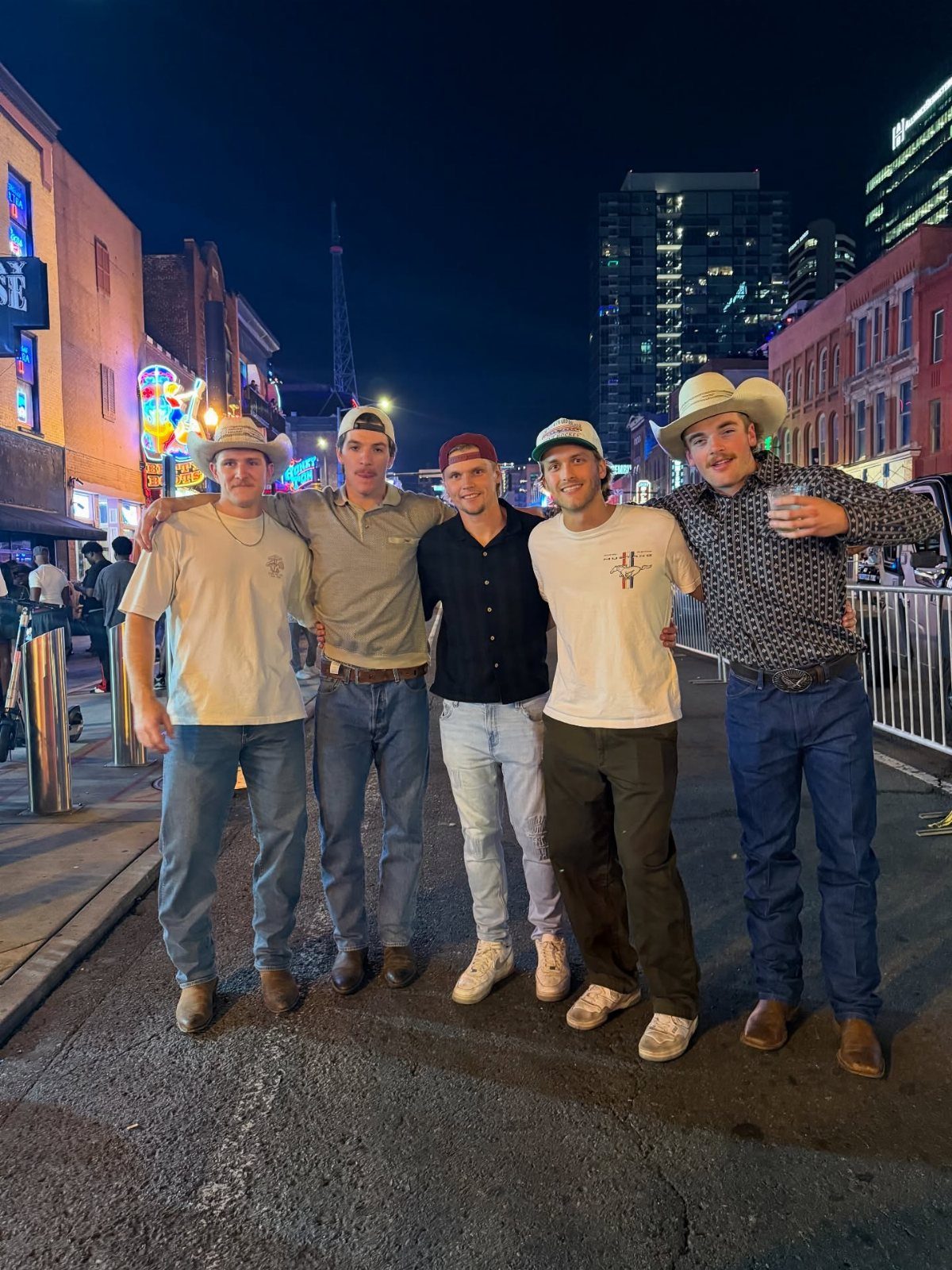 Five young men posing together on a city street at night wearing casual outfits and hats.