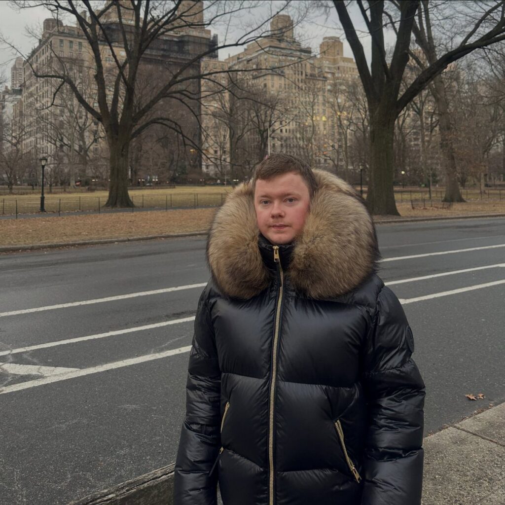 A young man wearing a black puffer jacket with a large fur hood standing outdoors.