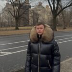 A young man wearing a black puffer jacket with a large fur hood standing outdoors.