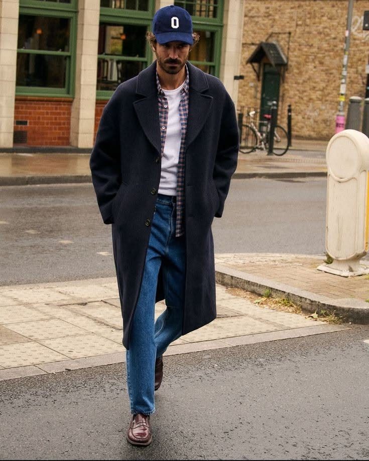 Man walking in a city wearing a long navy wool coat, plaid shirt, and blue jeans.