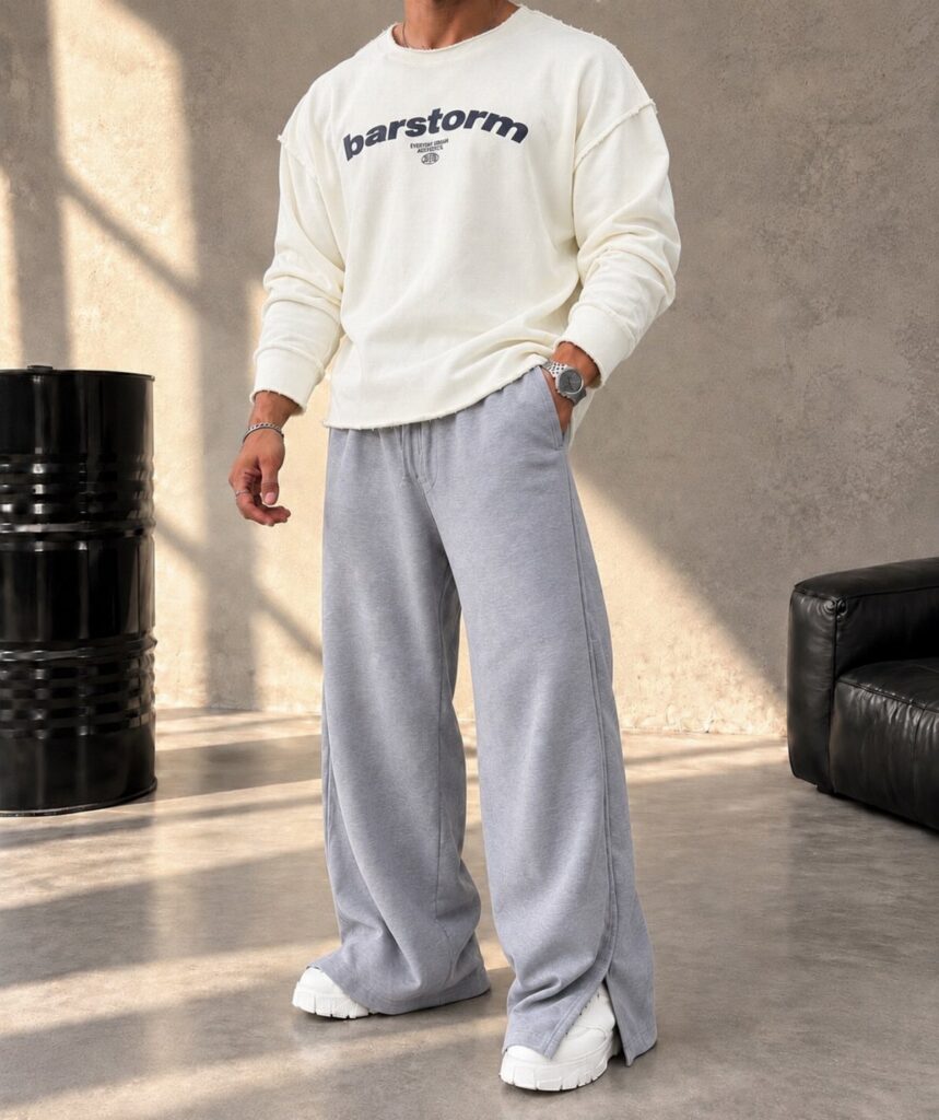 A person wearing a white sweatshirt, wide grey sweatpants, and white sneakers in a studio.