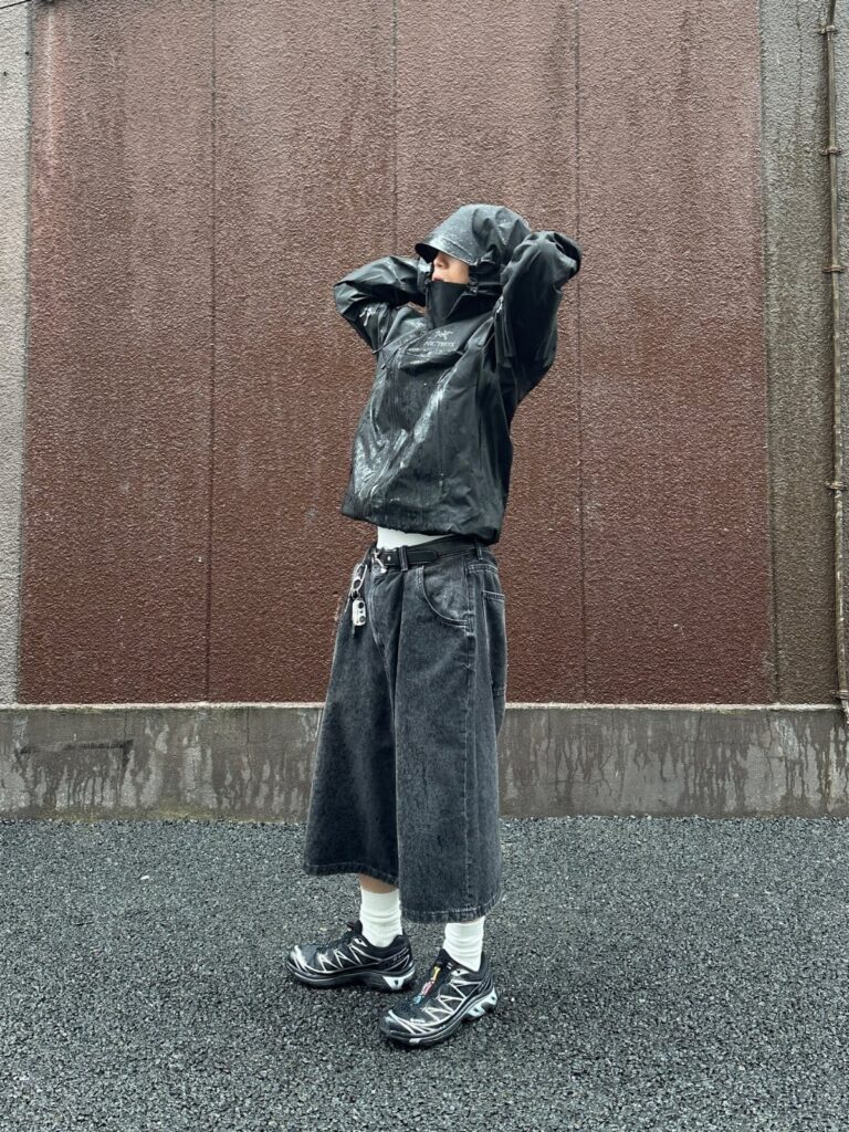 A person in a black hooded rain jacket and dark shorts standing against a brown wall.
