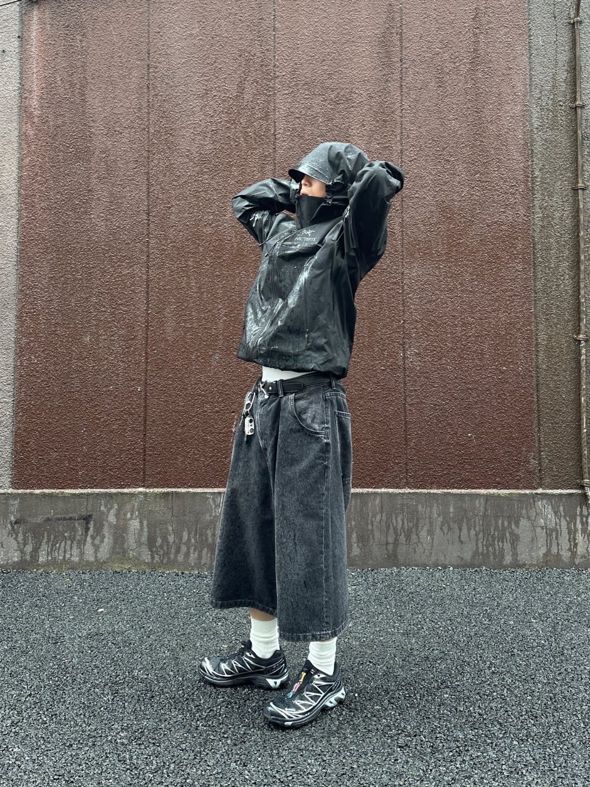 A person in a black hooded rain jacket and dark shorts standing against a brown wall.